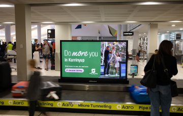 Airport baggage claim area with a luggage carousel in the foreground and travellers standing nearby. A large digital display on the carousel shows a green “Do more you in Karrinyup” 